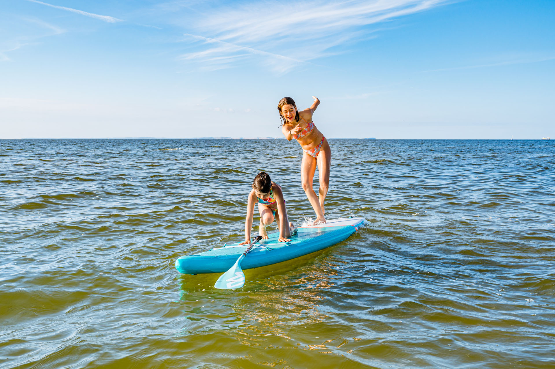 Two girls on an SUP on the water, the younger tries to get up. The older one stands and tries to balance. Only the water in the background